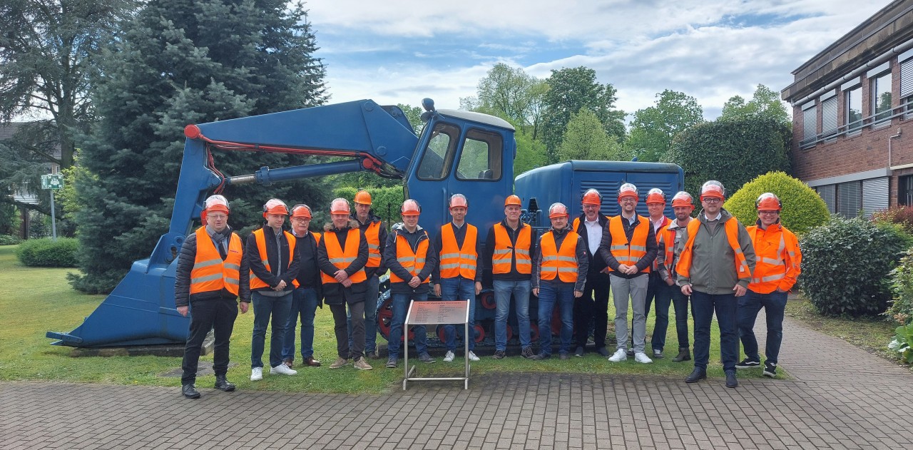 EMMEGI-Komatsu - EMMEGI GmbH Construction crew in orange safety vests and helmets posing in front of a large blue excavator outdoors. Group of workers on a site, wearing high-visibility jackets and hard hats.