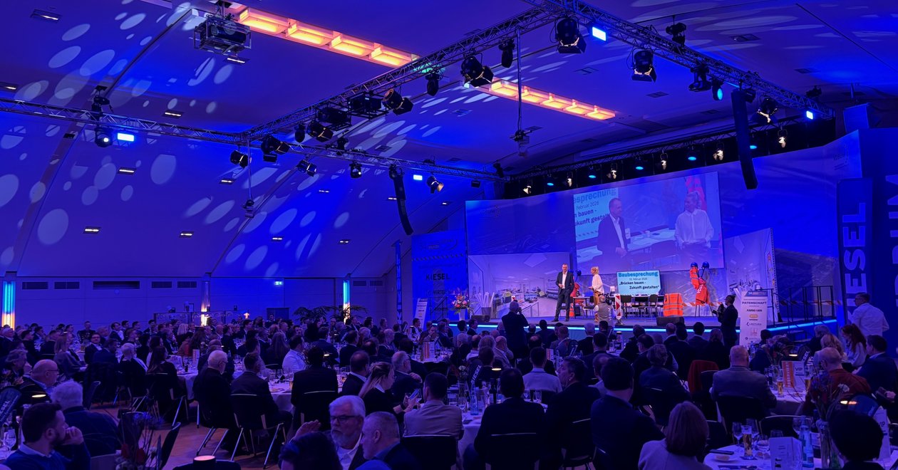 Large conference hall with a blue-lit stage, presenters on stage and a seated audience at round tables during a formal event.