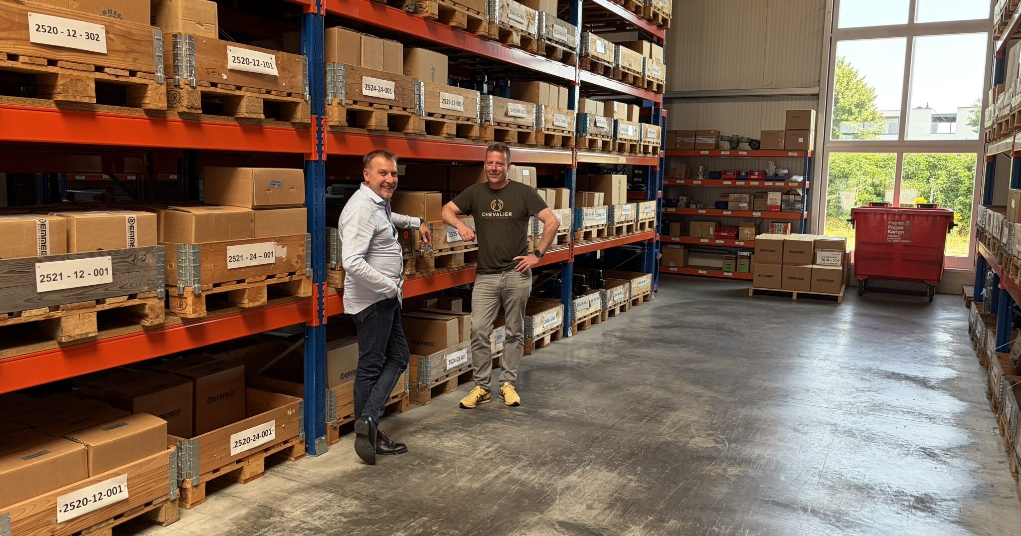 Two men stand in a warehouse aisle between tall orange-and-blue pallet racks loaded with boxes and crates.